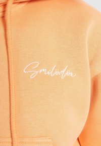 Sweat à capuche orange clair avec texte brodé en cursive blanche « Smilodox » sur la poitrine et détail cordon de serrage à la capuche.