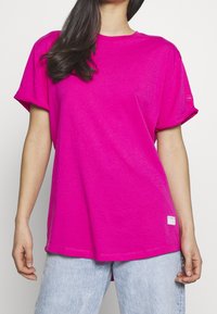 T-shirt en coton fuchsia avec des manches retroussées, col rond, coupe ample et une petite étiquette logo à l'ourlet. Associé à un jean bleu clair.