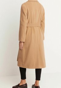 Camelfärgad kappa i mjukt tyg, med bälte i midjan, långa ärmar och sidfickor, tillsammans med svarta leggings och loafers.