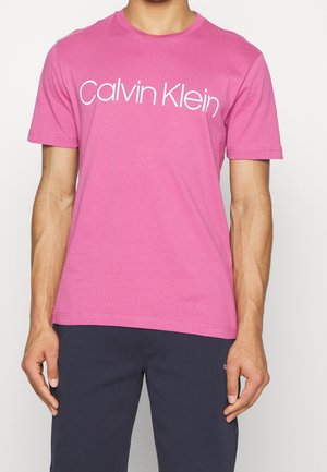 Mężczyzna ubrany w różowy t-shirt Calvin Klein z białym napisem logo, w zestawieniu z ciemnoniebieskimi spodniami, pokazany od szyi do połowy uda.