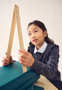 Righello di legno tenuto da un bambino, che misura una scatola di colore acqua su un tavolo di legno. Il bambino indossa un maglione grigio con volant e un colletto bianco.