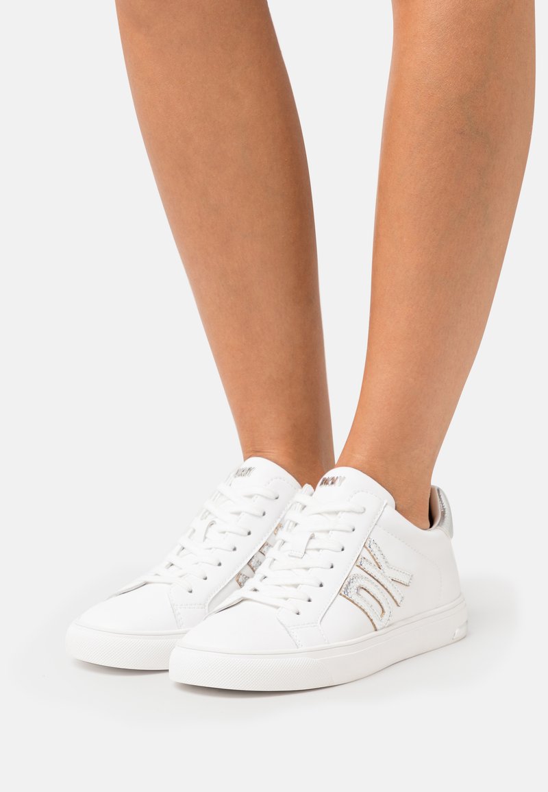 Baskets blanches avec des accents métalliques texturés, bout rond, semelle en caoutchouc et design à lacets. Présente le logo DK sur le côté et un col rembourré.