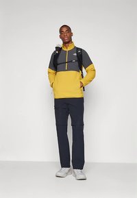 Mlad moški v rumeno-sivem zip-up jopiču, črnih hlačah, belih supergah in s črnim nahrbtnikom, ki stoji proti enobarvni ozadju.