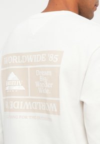 Λευκή μπλούζα με φούτερ που διαθέτει μια μεγάλη μπεζ γραφική εκτύπωση στην πλάτη, με το κείμενο "WORLDWIDE '85" και "Dream Big, Wander Wide." Περιλαμβάνεται εκτυπωμένο λογότυπο.