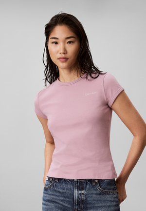 Młoda kobieta z mokrymi włosami, ubrana w dopasowany różowy t-shirt Calvin Klein i niebieskie dżinsy, na jednolitym szarym tle.
