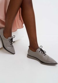 Chaussures en cuir gris à lacets avec un bout pointu, des lacets noirs contrastants, des détails perforés, et une semelle noire plate et fine.