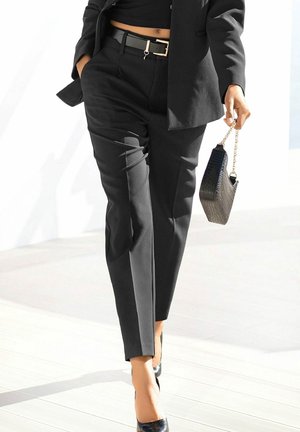 Personne portant un pantalon noir sur mesure, un top court noir et un blazer, tenant un petit sac à main noir avec une bandoulière en chaîne, marchant vers l'avant.