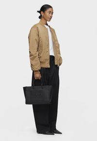 Czarna skórzana torba typu tote z wytłoczonym logo "TOUS", w połączeniu z beżową kurtką bomberką i luźnymi czarnymi spodniami. Gładka tekstura, strukturalny design.
