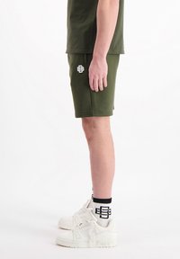 Groene katoenen shorts met een klein wit logo, gecombineerd met witte sneakers en zwart-wit gestreepte sokken. Zijaanzicht.