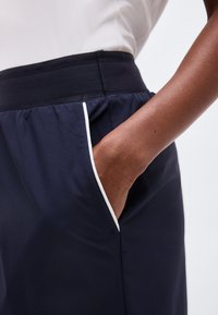 Χέρι ατόμου στην τσέπη σκούρου ναυτικομπλε παντελονιού με λευκή διακόσμηση, που φοράει λευκό πουκάμισο, πάνω σε απλό ανοιχτό φόντο.