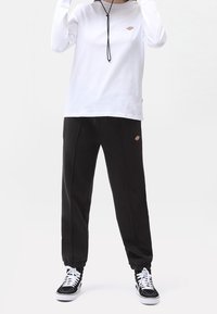 White long-sleeve shirt with a logo on the chest, paired with black tapered sweatpants featuring elastic cuffs and two front seams. Black sneakers.