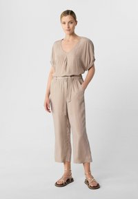 Beige Jumpsuit mit V-Ausschnitt, kurzen Ärmeln, Taillengebund und verkürzten Beinen, kombiniert mit Riemchensandalen mit flacher Sohle.