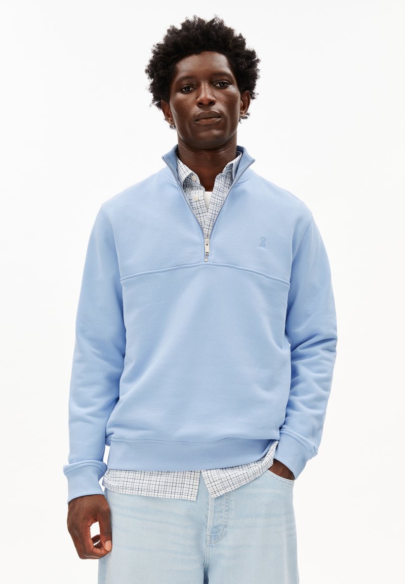 Homme portant un sweat à col zippé bleu clair sur une chemise blanche à carreaux et un jean en denim clair, debout devant un fond blanc uni.
