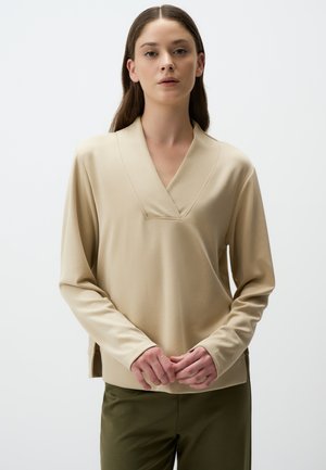 Beige long-sleeve top with a V-neck design and textured detailing at the neckline, paired with olive-green pants. Smooth fabric.