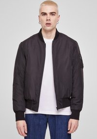 Chaqueta bomber negra con cremallera frontal, puños y dobladillo acanalados, con un bolsillo lateral. Llevada sobre una camiseta blanca y jeans azules.