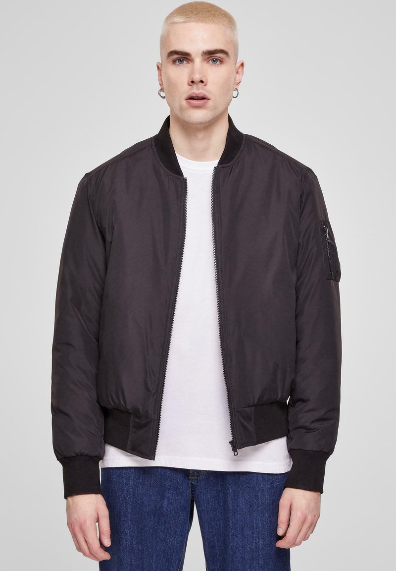 Chaqueta bomber negra con cremallera frontal, puños y dobladillo acanalados, con un bolsillo lateral. Llevada sobre una camiseta blanca y jeans azules.