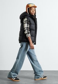 Gilet en duvet noir sur une chemise à carreaux, jeans évasés bleu clair, et baskets noires avec des rayures blanches. Casquette marron et bleue avec des rabats pour les oreilles.