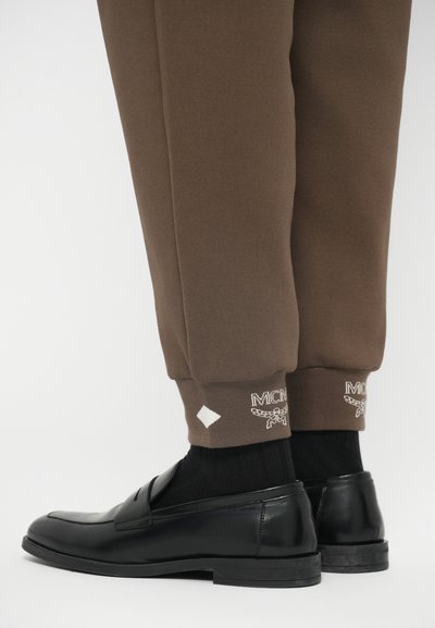 Onderbenen in bruine joggers met witte logo-borduursel, gecombineerd met zwarte loafers en zwarte geribde sokken tegen een witte achtergrond.