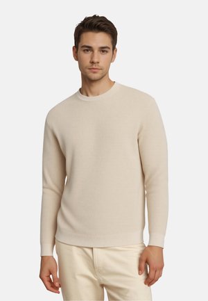 Pull côtelé beige avec un col rond et des manches longues, présentant une texture douce et des poignets ajustés, porté avec un pantalon de couleur claire.