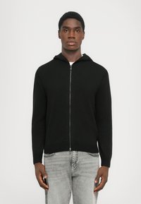Sort zip-up hættetrøje med en struktureret stof, der har hætte og ribbet kant. Modellen har lys grå jeans på og står foran en ensfarvet baggrund.
