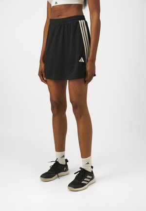 Kvinde iført sort Adidas sportskørt med hvide striber, sorte Adidas sneakers og hvide Adidas sokker, der står på en ensfarvet hvid baggrund.