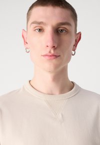 Krémová mikina s rib výstřihem a křížovým prošíváním. Modelka má na levém uchu stříbrný kruhový náušník.