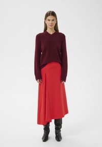 Bordeauxroter, gestrickter Pullover mit V-Ausschnitt und langen Ärmeln, kombiniert mit einem hochtaillierten, roten Rock mit Frontdrapierung und schwarzen kniehohen Stiefeln.