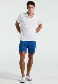 Polo shirt blanc à manches courtes avec de fines rayures horizontales, associé à un short bleu. Présente un petit logo sur le shirt et des baskets blanches.