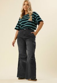 Mustat cargo-housut leveillä lahkeilla, joissa on kaksi sivutaskua ja pehmeä tekstuuri. Yhdistettynä vihreään ja mustaan raidoitettuun ylisuurikokoiseen t-paitaan, jossa on karhukuva.