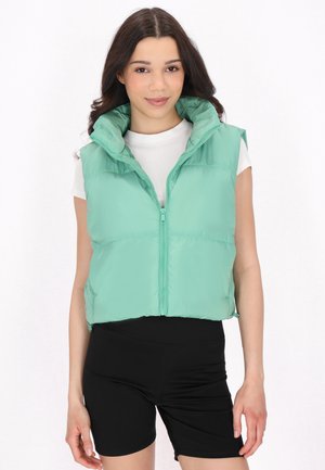 Mintgrøn puffervest med høj krave, lynlås og cropped design. Båret over en hvid skjorte og parret med sorte shorts.
