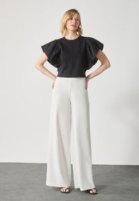 Black cropped top with wide, ruffled sleeves paired with high-waisted, wide-leg white trousers. Smooth fabric with a modern silhouette.