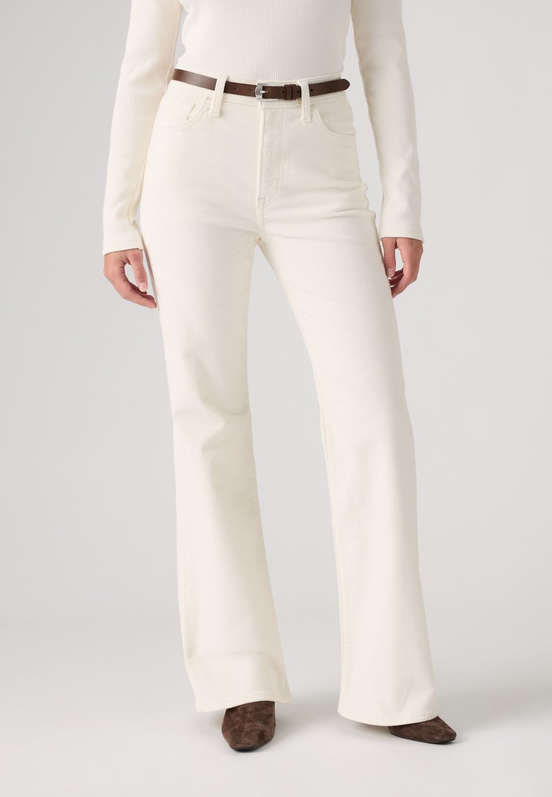 Personne portant un pantalon large taille haute écru, une ceinture marron, un haut côtelé écru assorti et des chaussures pointues marron sur un fond uni.