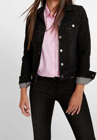 Černá džínsová bunda se stříbrnými knoflíky, předními kapsami a ohrnutými šedými manžetami, nošená přes světle růžovou košili, která předvádí neformální vrstvený vzhled.