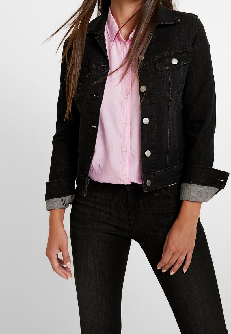 Černá džínsová bunda se stříbrnými knoflíky, předními kapsami a ohrnutými šedými manžetami, nošená přes světle růžovou košili, která předvádí neformální vrstvený vzhled.