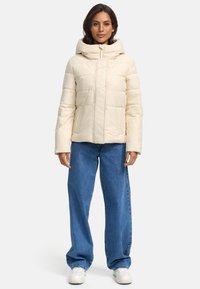 Doudoune crème avec capuche, présentant un design matelassé, une fermeture éclair à l'avant et des poches latérales. Associée à un jean bleu à jambes larges et des baskets blanches.
