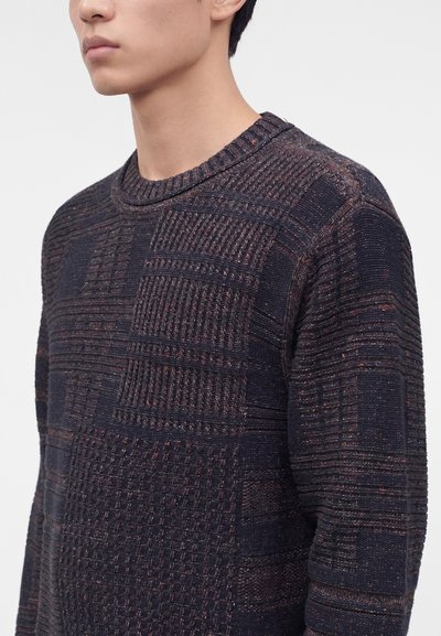 Jeune personne portant un pull en maille texturée foncée avec un motif patchwork et un col rond, montré de la poitrine au menton.