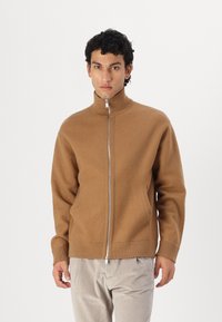 Beige jakke med glidelås og krage, laget av mykt materiale, med lange ermer og sidelommer, kombinert med lyse bukser.