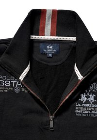 Černá mikina na zip s vysokým límcem, s límcem s bílými a červenými pruhy. Na přední straně vyšívaný grafický text a loga.