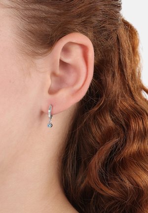 Orecchini a cerchio in argento con un piccolo pendente di gemma azzurra, caratterizzati da una texture liscia e un design semplice, indossati all'orecchio di una persona.
