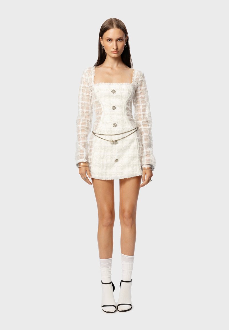 Weißes, strukturiertes Minikleid mit quadratischem Ausschnitt, durchsichtigen langen Ärmeln und dekorativen Knöpfen. Kombiniert mit weißen Socken und Absätzen mit schwarzen Akzenten.