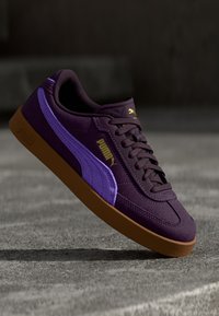 Ljubičaste suede tenisice s svijetloljubičastom prugom, zlatnim PUMA logotipom i smešnim gumenim potplatom. Klasični dizajn s teksturiranim detaljima na gornjem dijelu.