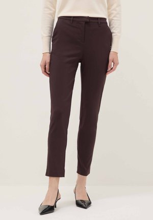 Vrouw draagt strakke donkerbruine enkellange broek, crème lange mouwen top en zwarte puntige hakken, staand tegen een effen achtergrond.