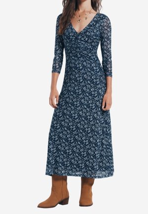 Mujer con un vestido midi de manga larga azul marino con estampado de cachemira y escote en v, y botas de ante marrón hasta la mitad de la pantorrilla, posando frente a un fondo blanco.