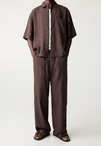 Mężczyzna w luźnej, brązowej lnianej koszuli z krótkim rękawem zapinanej na guziki oraz dopasowanych szerokich spodniach w tym samym odcieniu, z ciemnobrązowymi sandałami.