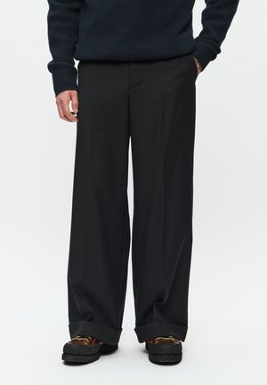 Pantalons larges noirs en tissu lisse, avec des plis avant, une coupe ajustée et des poignets légèrement retroussés, associés à des chaussures sombres.
