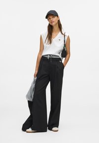 Γυναίκα με λευκό άκοπο τοπ, μαύρο παντελόνι με φαρδιά πόδια, μαύρο καπέλο, λευκά αθλητικά παπούτσια, κρατώντας ένα γκρι ρούχο και μια μικρή μαύρη τσάντα.