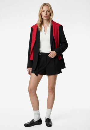 Lyshåret kvinde iført sort blazer og shorts, hvid ribbet top, rød sweater hængende over skuldrene, hvide sokker og sorte loafers, stående.