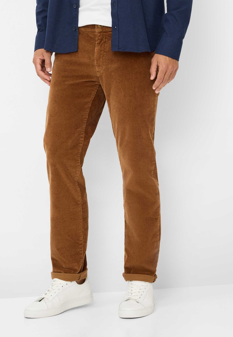 Braune Baumwollcord-Hose mit geradem Schnitt, hochgekrempelten Säumen und strukturierter Oberfläche. Kombiniert mit weißen Sneakern und einem navyblauen Hemd mit Knöpfen.