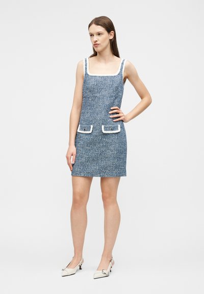 Femme debout, main sur la hanche, portant une mini robe sans manches en tweed bleu avec des poches bordées de blanc et des chaussures blanches à talons bas.