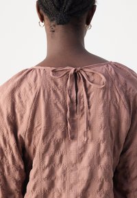 Vue de dos d'une personne aux cheveux courts tressés, portant des boucles d'oreilles anneaux dorés et un chemisier mauve texturé noué d'un nœud au col.